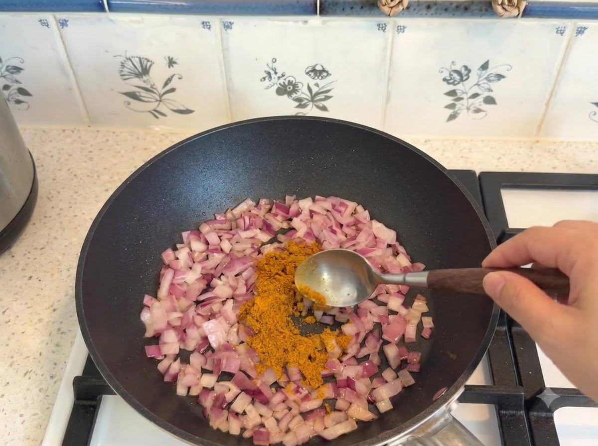 Currypulver wird mit einem Löffel abgemessen und zu den angebratenen Zwiebeln in der Pfanne gegeben.