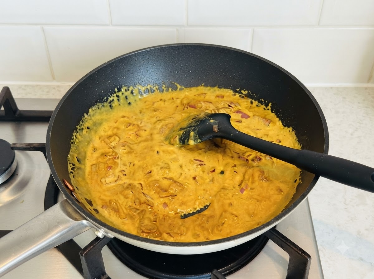 Reichhaltige gelbe Currysauce wird in einer dunklen Pfanne mit einem schwarzen Silikonspatel umgerührt.