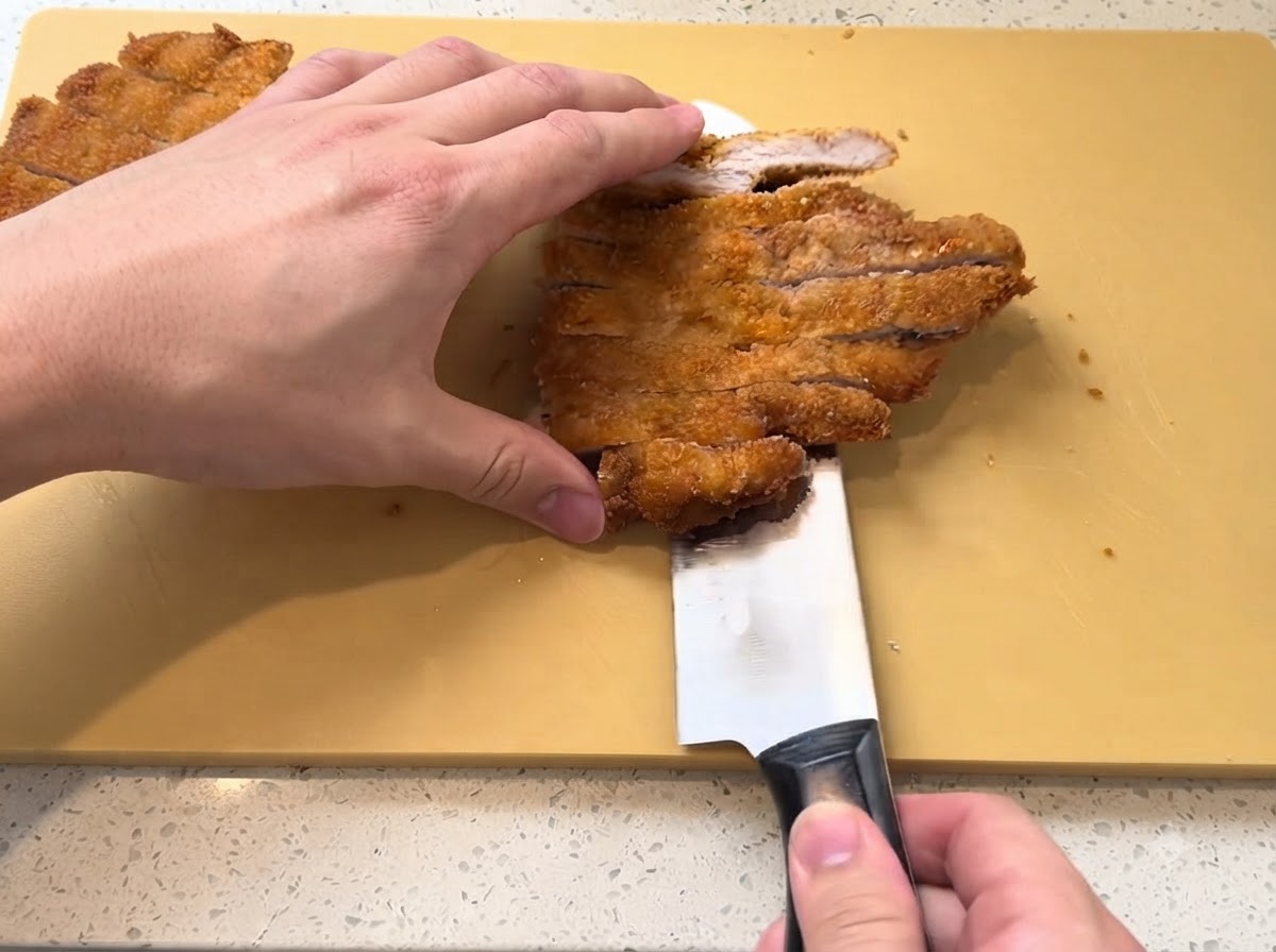 Ein goldbraun frittiertes Schweineschnitzel wird mit einem scharfen Messer auf einem gelben Schneidebrett in Streifen geschnitten.