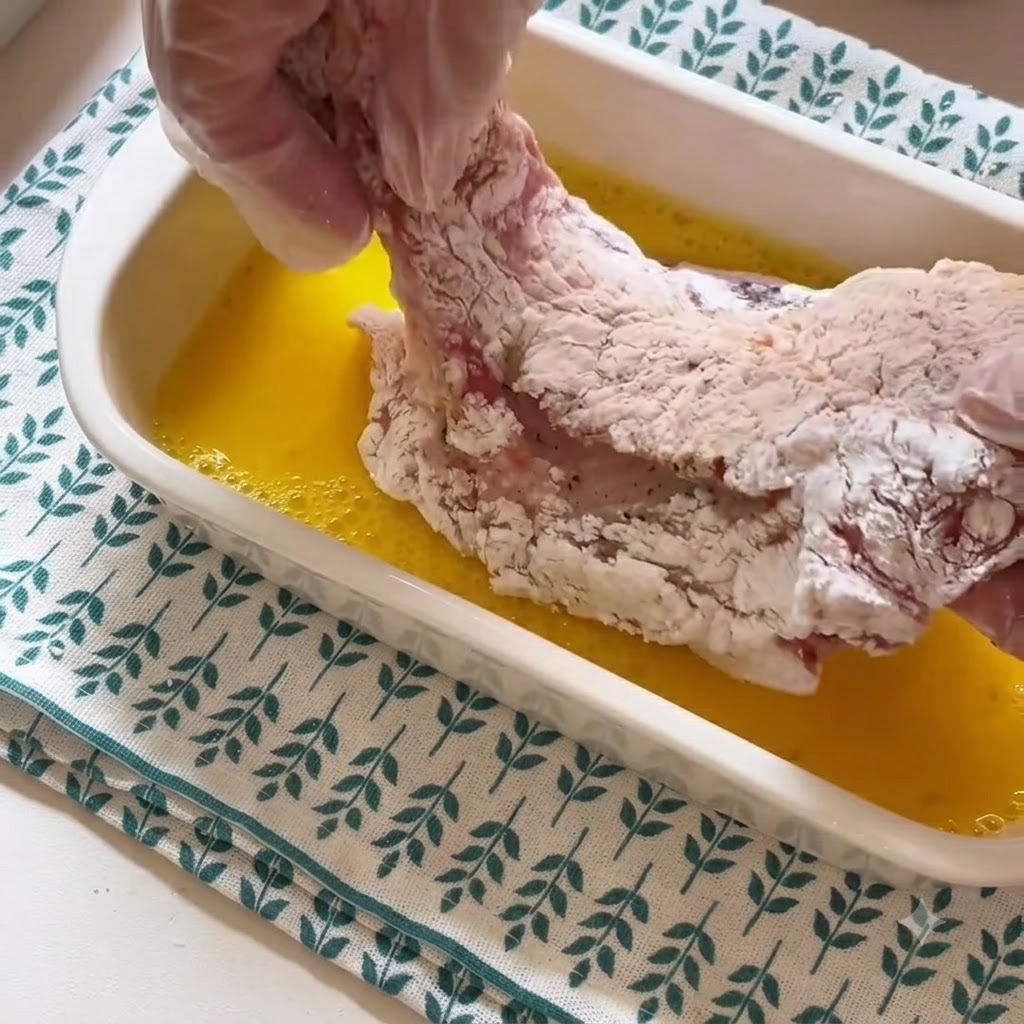 Eine behandschuhte Hand hebt ein mit Stärke überzogenes Schweinekotlett aus einem Behälter mit gelber, geschlagener Eierflüssigkeit.