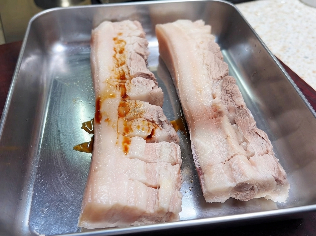 Zwei Stücke eingeritzter Schweinebauch auf einem Metallblech, über die dunkle Sojasauce gegossen wird.