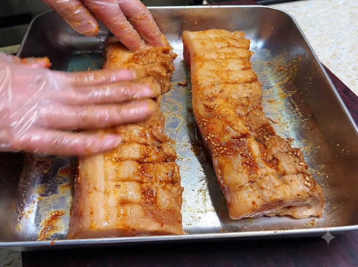 Behandschuhte Hände massieren dunkle Marinade und trockene Gewürze in die eingeritzte Oberfläche zweier Schweinebauchstreifen.