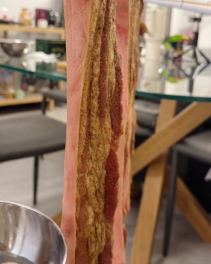 Schweinebauch hängt vertikal an einem Gestell, während die Haut mit Knusperhautwasser bestrichen wird.