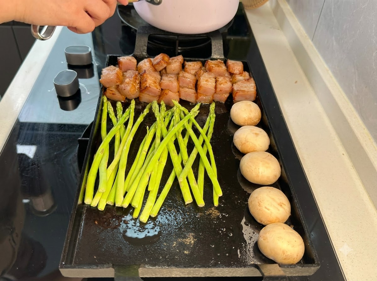 Frische grüne Spargelstangen liegen auf der Grillpfanne neben Fleisch und Pilzen.