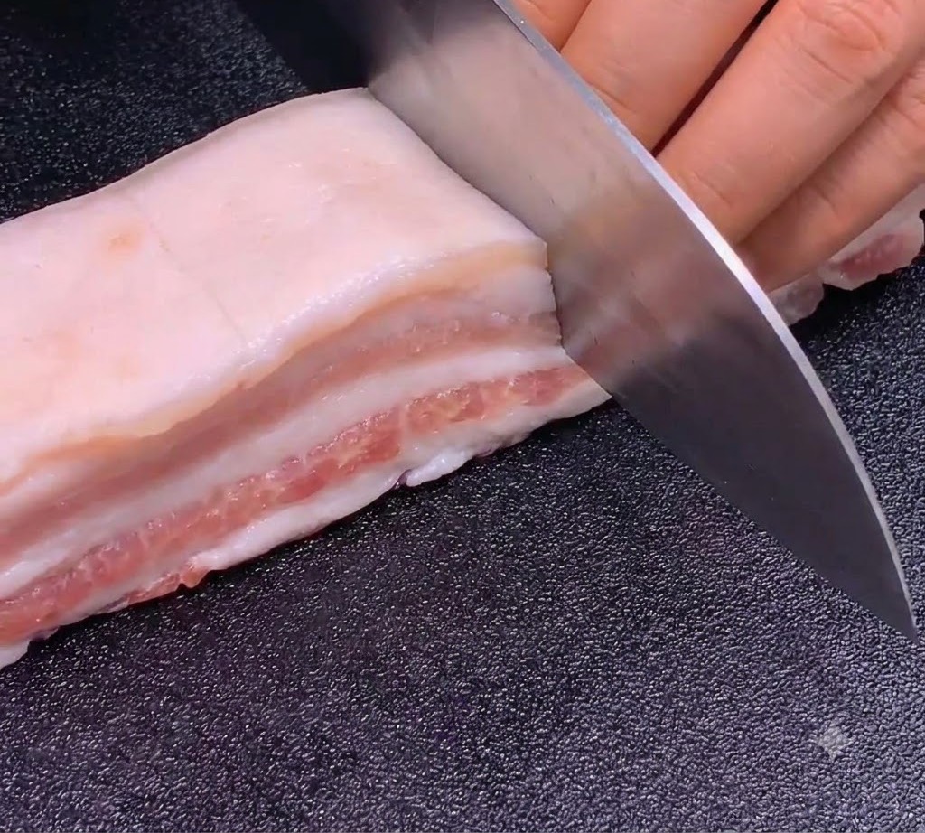 Ein scharfes Messer schneidet präzise und gleichmäßig in einen Schweinebauchstreifen auf einem dunklen Schneidebrett.