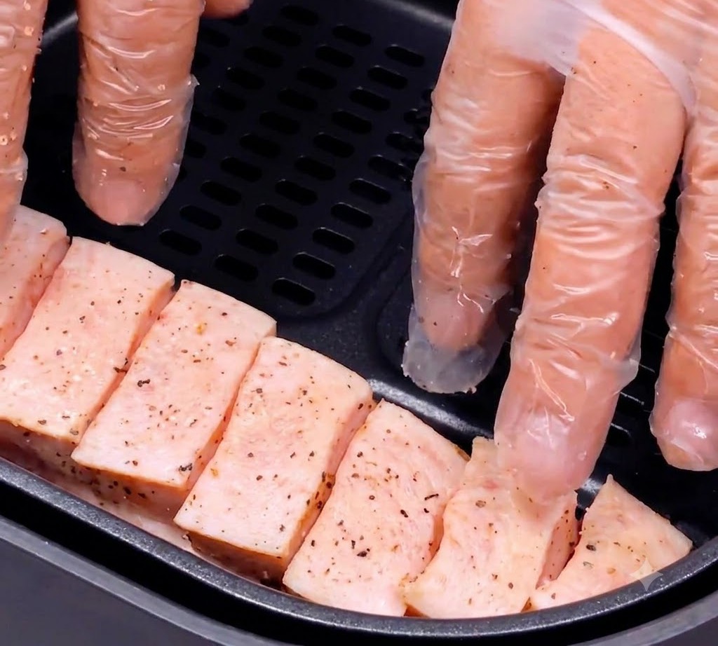 Gewürzte Schweinebauchstreifen werden von handschuhbedeckten Händen sorgfältig in den dunklen Korb einer Heißluftfritteuse gelegt.