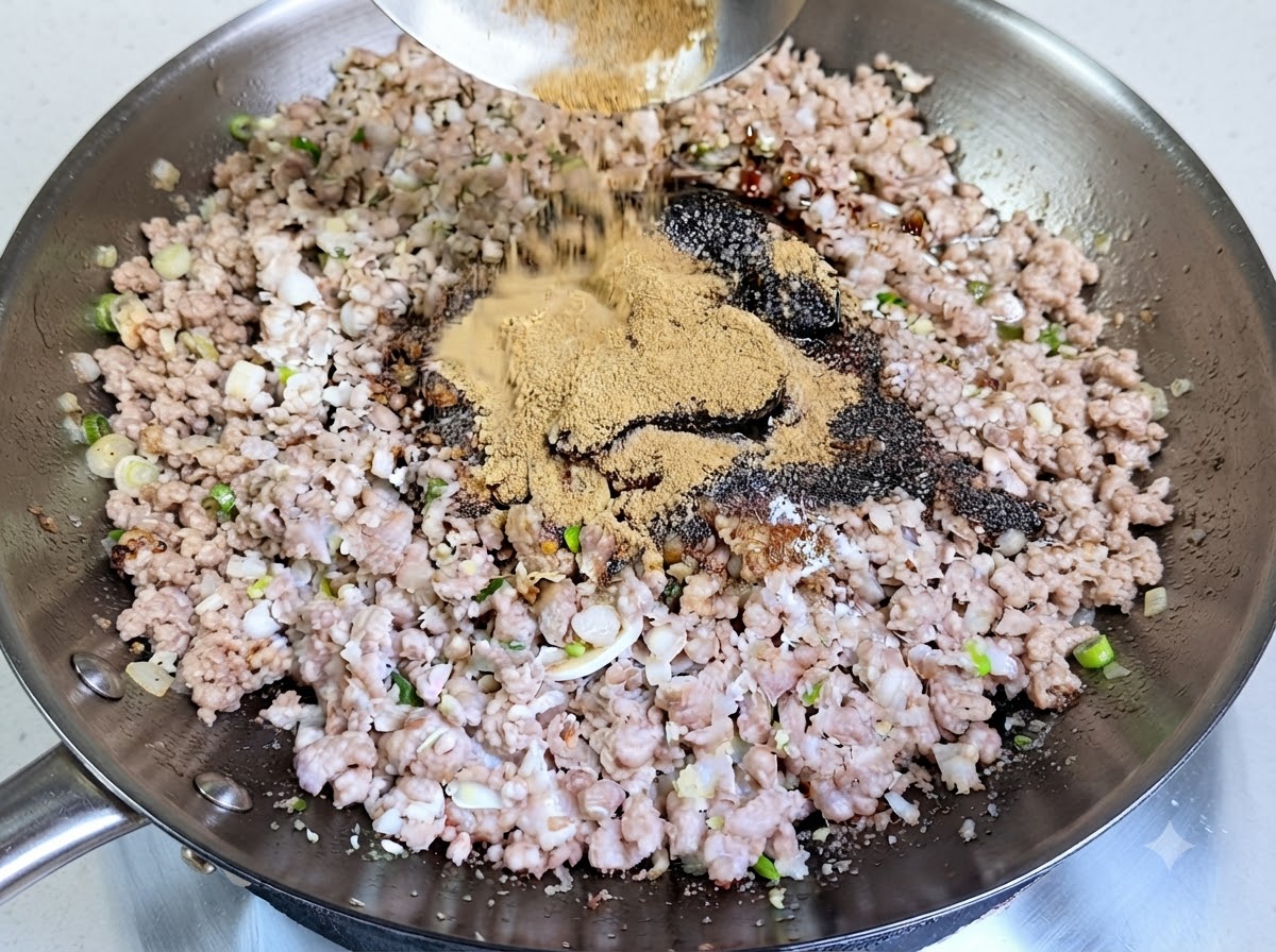 Ein großzügiger Haufen hellbraunem Gewürzpulver wird aus einer Kelle in den Wok über das gewürzte Schweinehackfleisch gegeben.