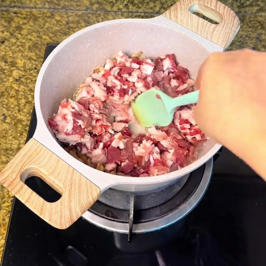 Gewürfeltes Schweinefleisch wird in den Topf mit dem ausgelassenen Fett gegeben und mit einem grünen Spatel verrührt.