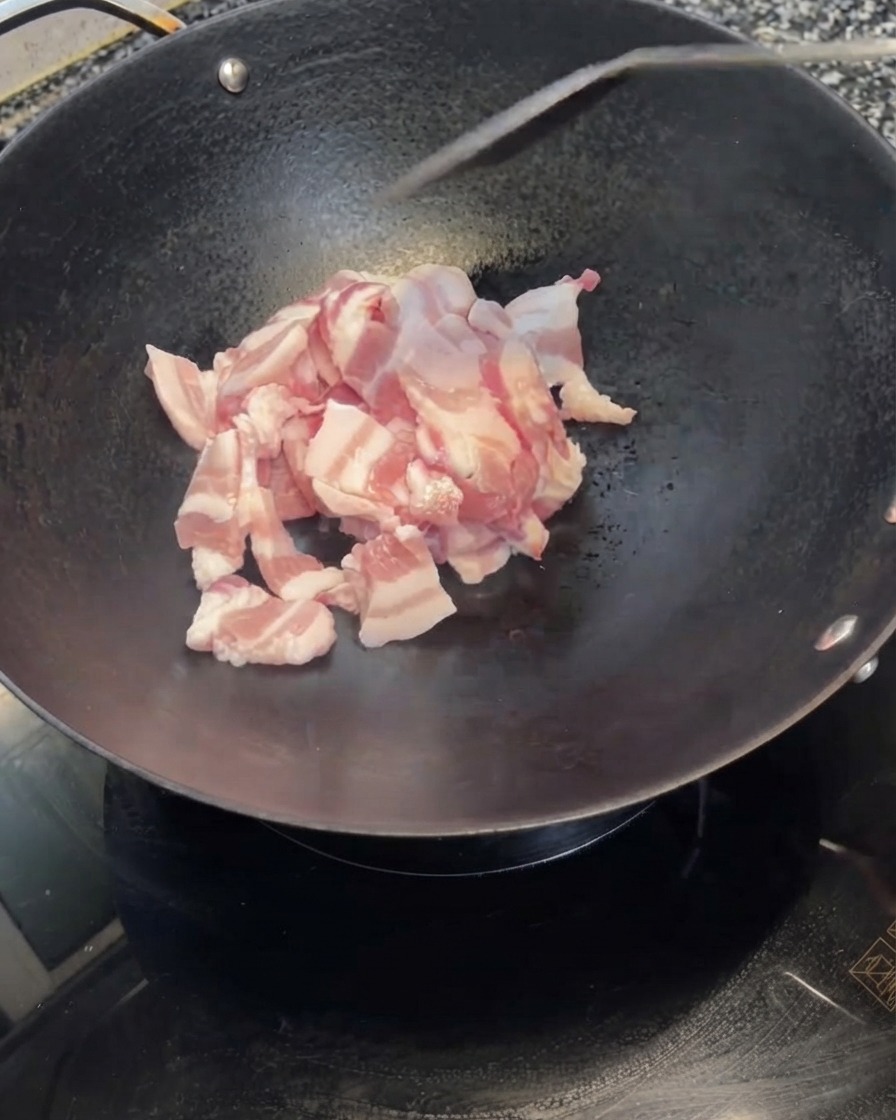 In Scheiben geschnittener Schweinebauch wird aus einer weißen Schüssel in einen heißen schwarzen Wok geschüttet.