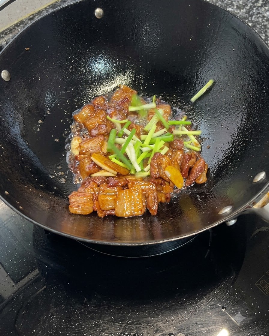 Frische Frühlingszwiebelstücke werden über karamellisierenden Schweinebauchstücken in einem Wok verstreut.