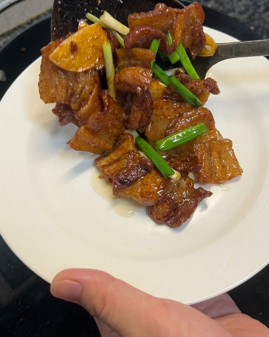 Gekochter Sojasaucen-Schweinebauch wird mit einem Pfannenwender aus einem Wok auf einen sauberen weißen Teller gehoben.