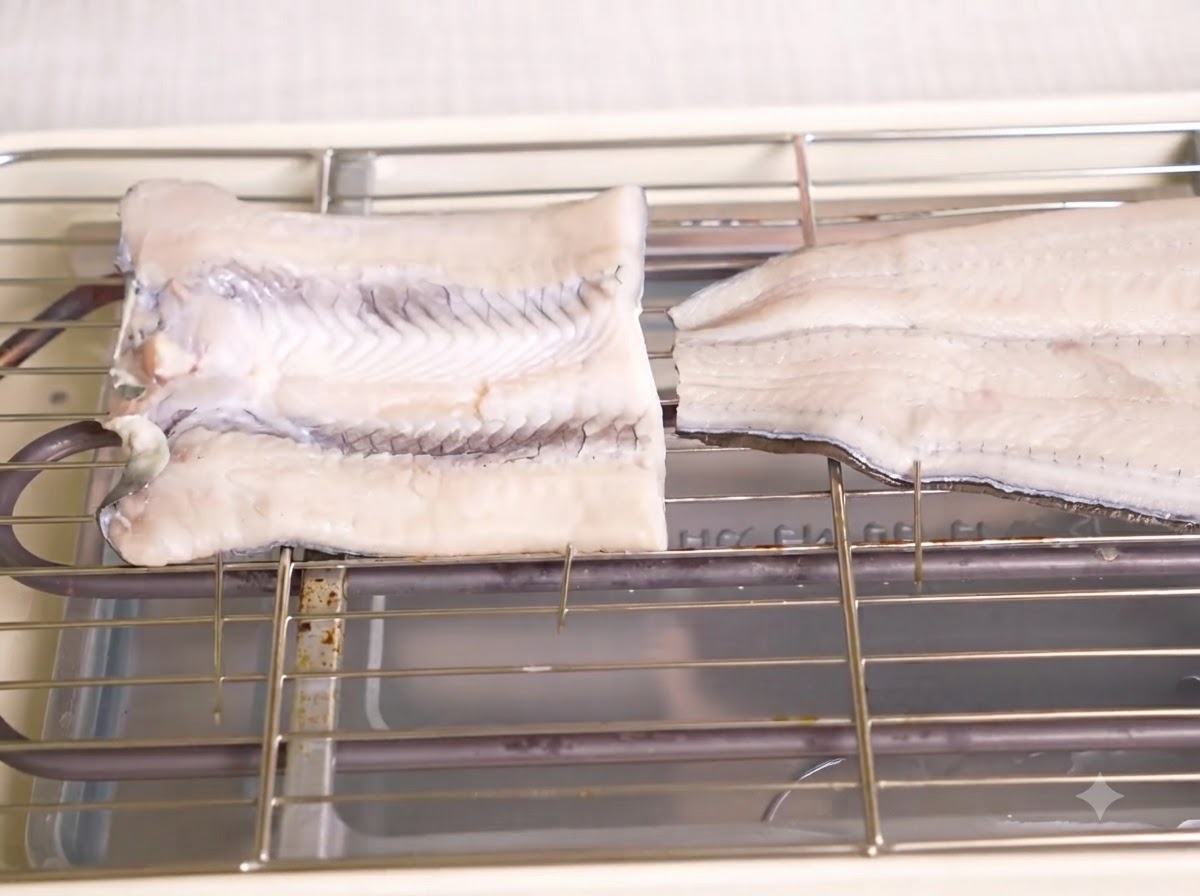 Zwei Stücke aufgespießter, roher Aal auf einem Metallrost über einem Elektrogrill.