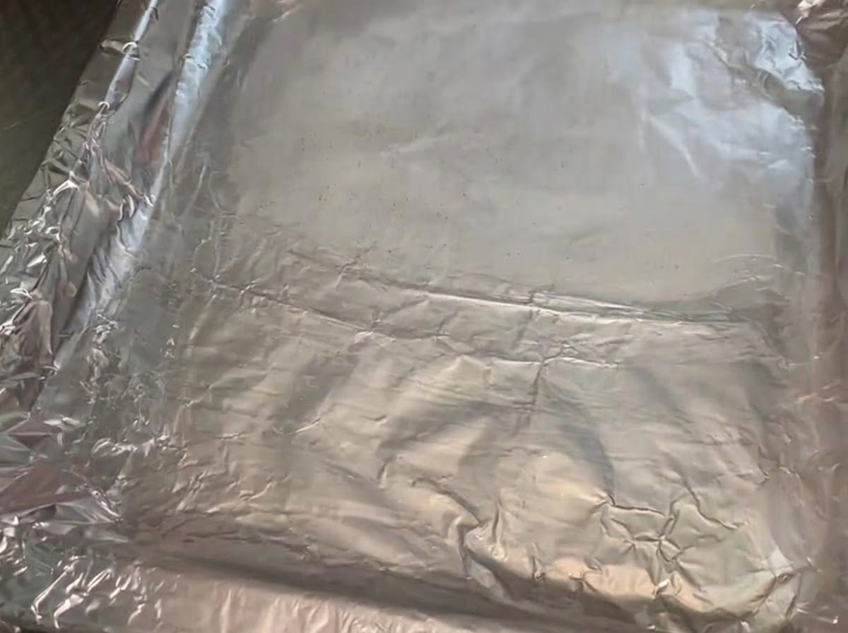 Ein Metallbackblech, ausgelegt mit zerknüllter Alufolie, die mit einer glänzenden Schicht Speiseöl bestrichen wurde.