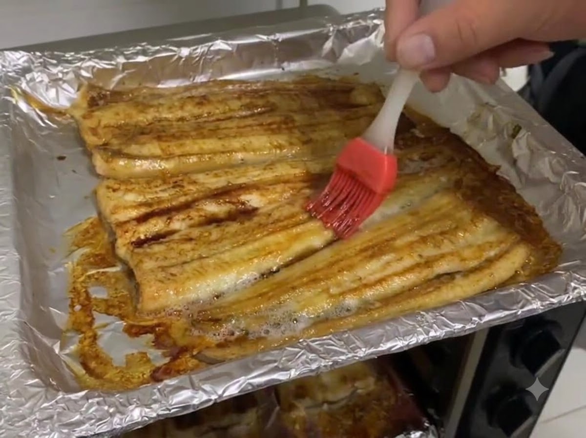 Dunkle Sojasauce wird auf die Oberfläche von gebackenen Aalfilets auf einem mit Alufolie ausgelegten Backblech gestrichen.