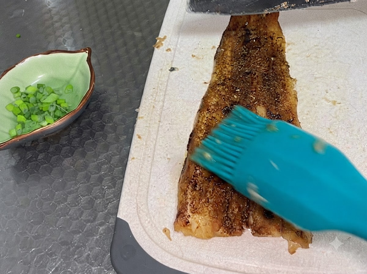 Ein fertig gebackenes Aalfilet wird auf einem weißen Schneidebrett neben einer kleinen grünen Schüssel mit gehackten Frühlingszwiebeln in Scheiben geschnitten.