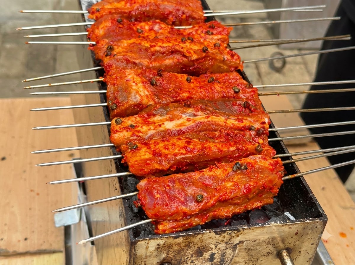 Mehrere Spieße mit rot überzogenen Rippchen sind aufgereiht und grillen auf einer Holzkohlegrillstelle.