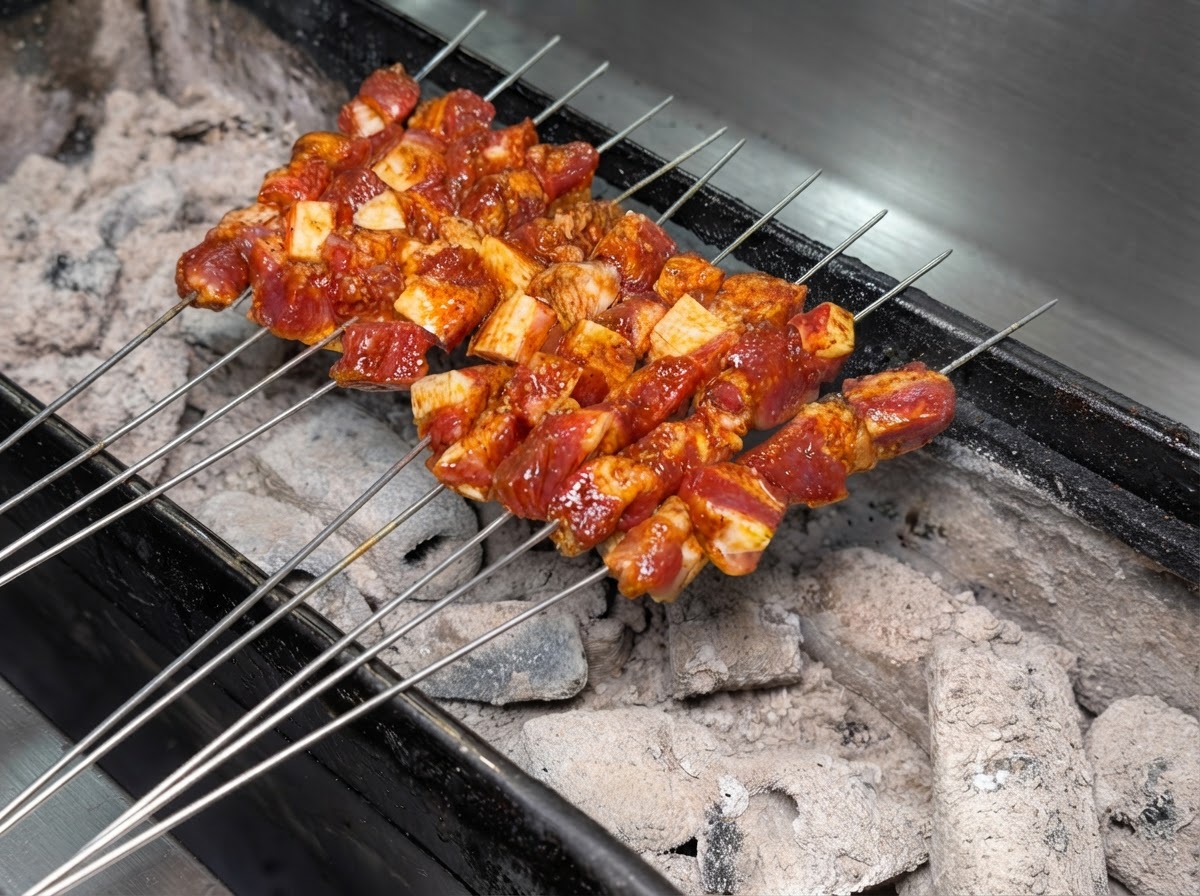 Mehrere Metallspieße mit gewürztem Schweinefleisch liegen ordentlich auf einem brennenden Holzkohlegrill.