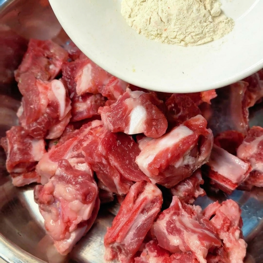 Gewürze und flüssige Würzmittel werden direkt auf rohe Schweinerippchen in einer Rührschüssel aus Metall gegossen.