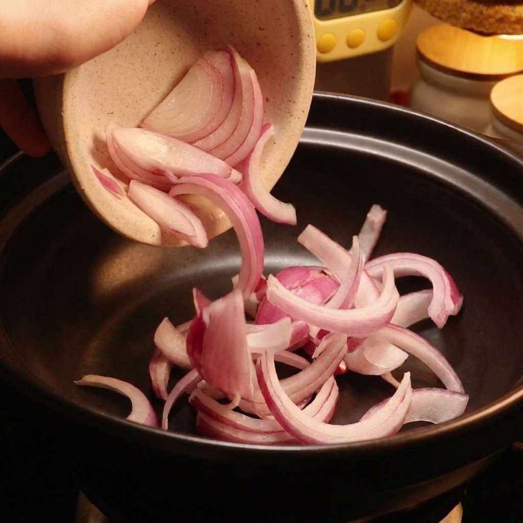 Rote Zwiebelscheiben werden aus einer Keramikschüssel in einen schwarzen Topf geleert.