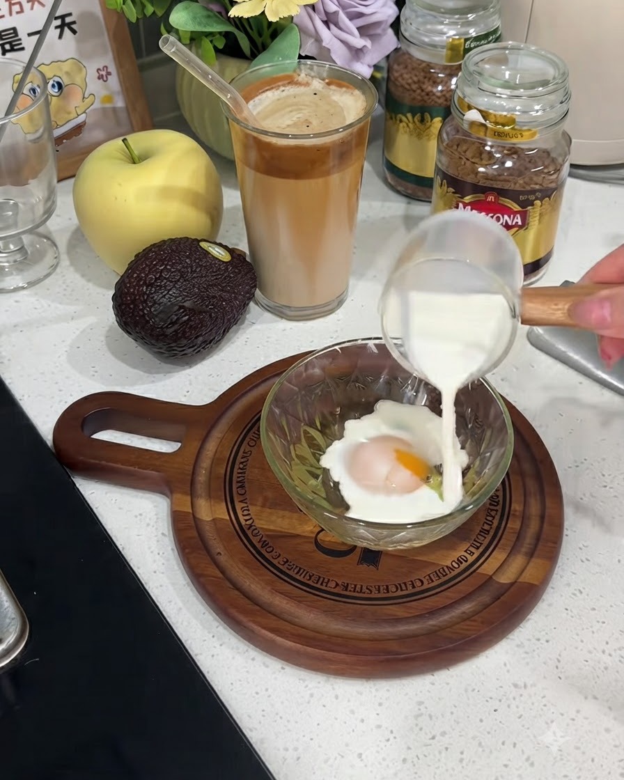 Milch wird aus einer kleinen Glaskanne in eine Schüssel mit einem aufgeschlagenen Ei gegossen.