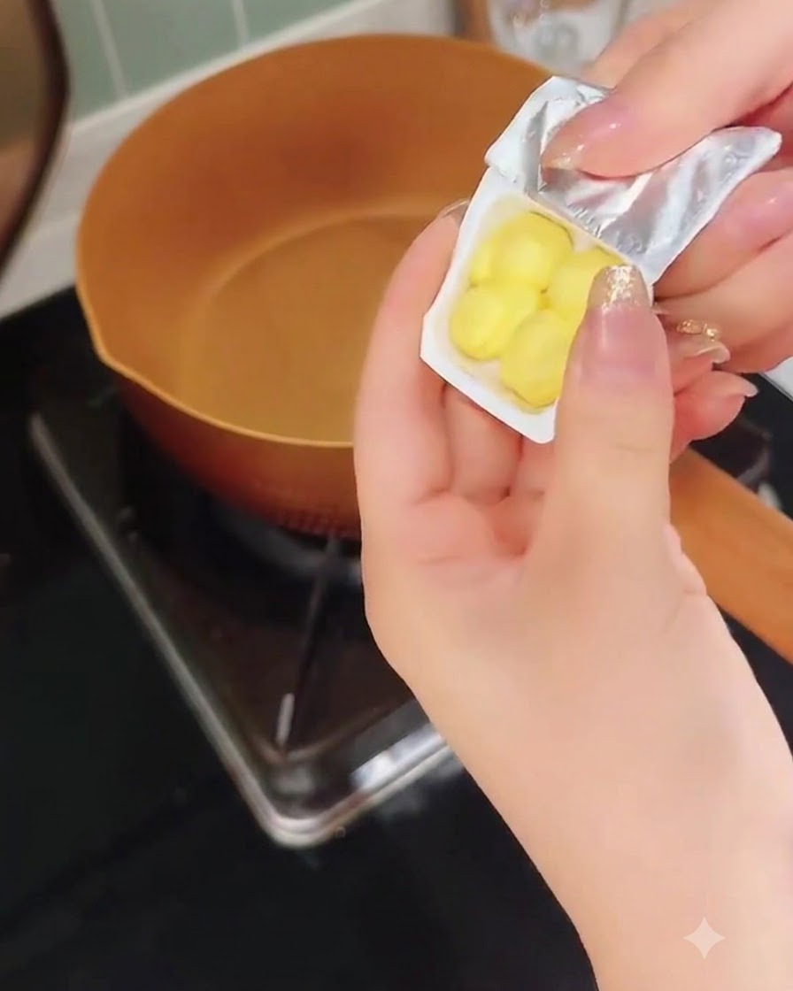 Eine Hand schält eine kleine Plastikverpackung mit gelben Butterstücken über einer warmen Kupferpfanne aus.