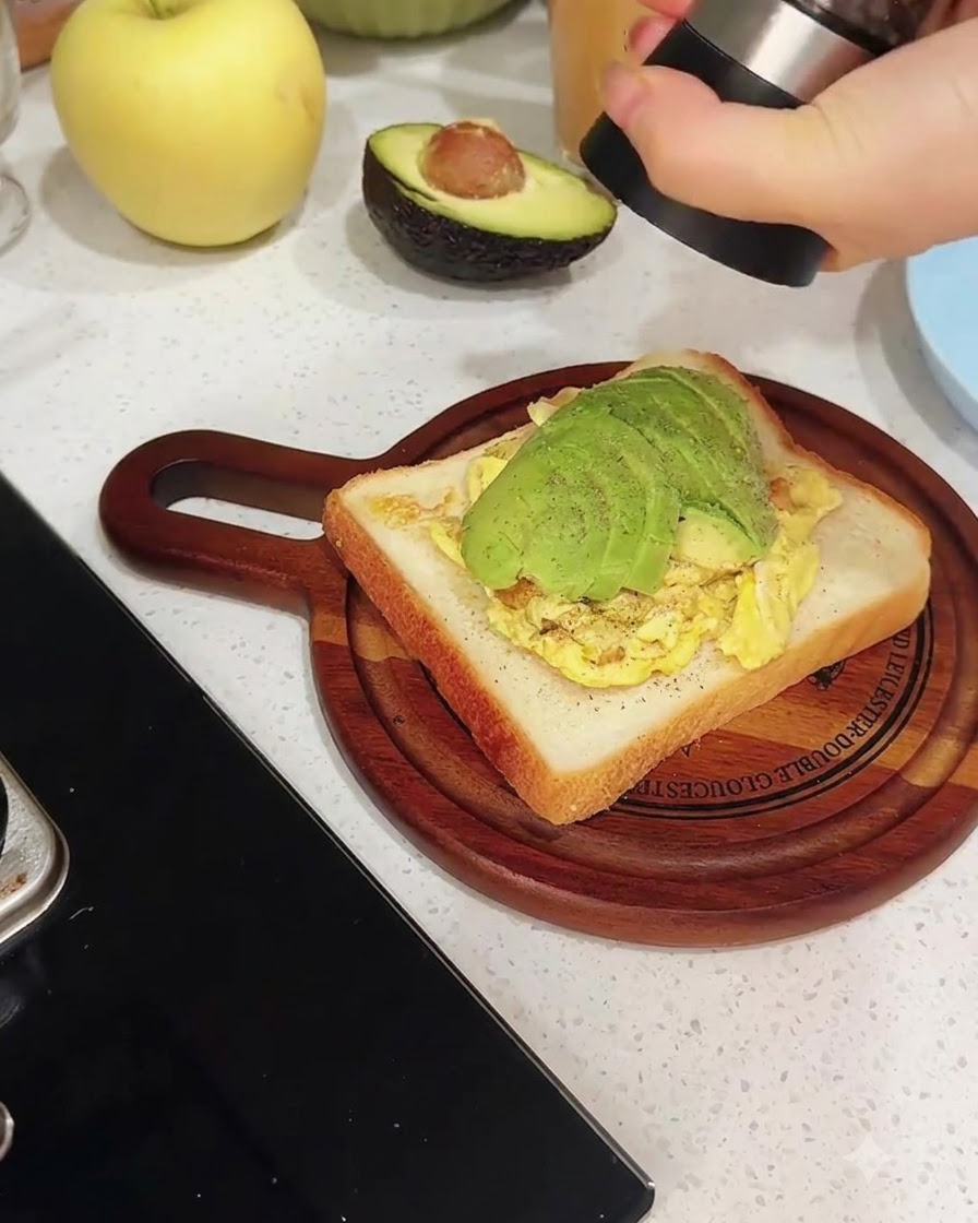 Eine Pfeffermühle mahlt frischen schwarzen Pfeffer direkt über eine Toastscheibe, die mit Rührei und Avocadoscheiben belegt ist.