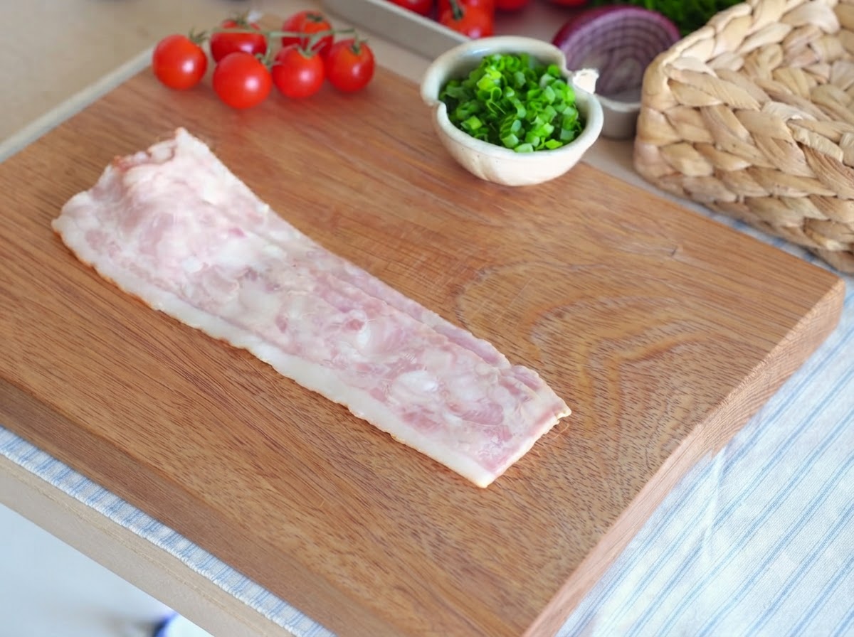 Ein einzelner roher Speckstreifen liegt auf einem Holzschneidebrett und ist bereit zum Schneiden.