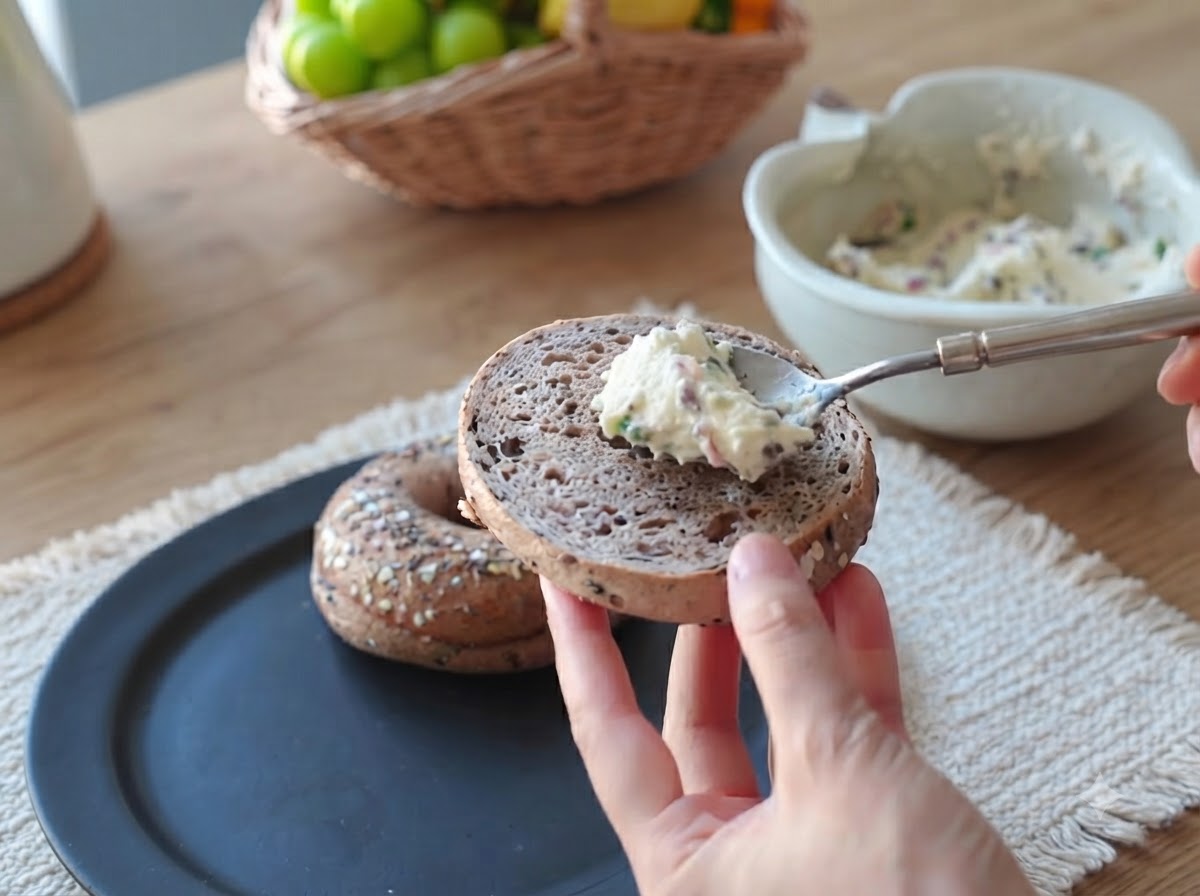Ein Löffel cremiger Bacon-Frühlingszwiebel-Aufstrich wird auf die Oberfläche einer Bagelhälfte gestrichen.