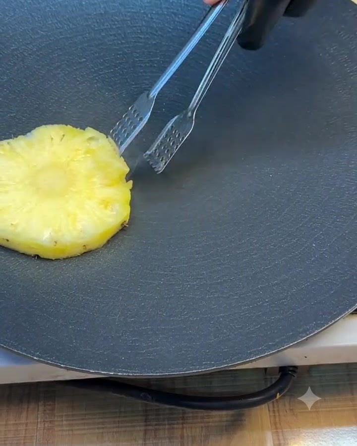 Ein einzelner Ananasring wird auf einer dunklen Grillplatte mit einer Metallzange gewendet.