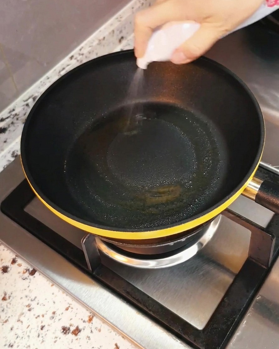 Eine Hand sprüht Speiseöl gleichmäßig in eine schwarze Bratpfanne auf einer Herdstelle.