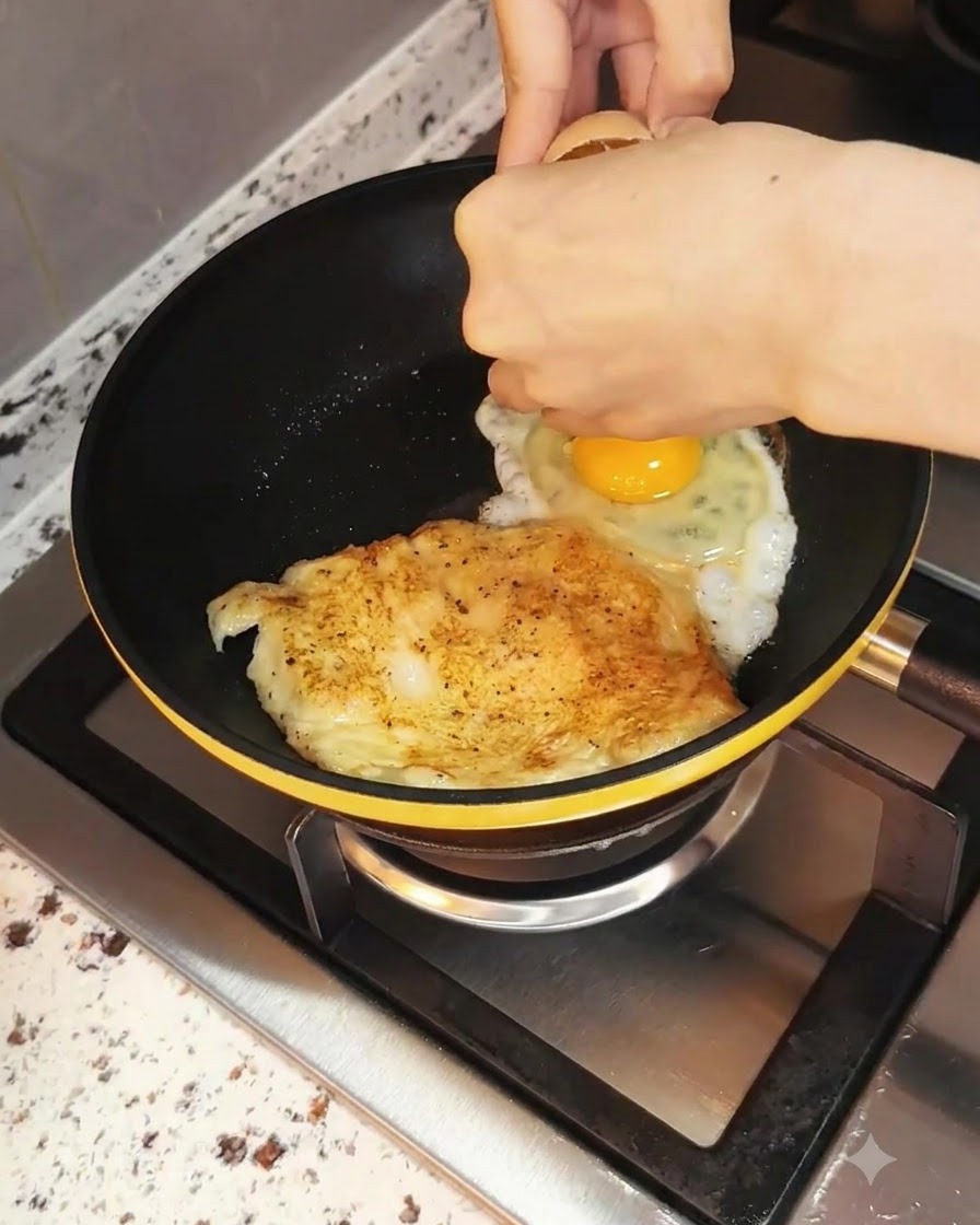 Ein Ei wird neben der bratenden Hähnchenbrust in die Bratpfanne geschlagen.