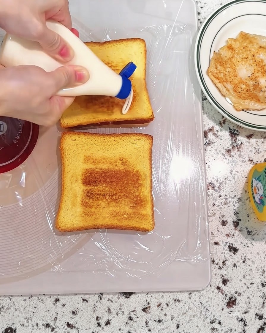 Mayonnaise wird aus einer Flasche auf getoastete Brotscheiben auf einem Schneidebrett gedrückt.