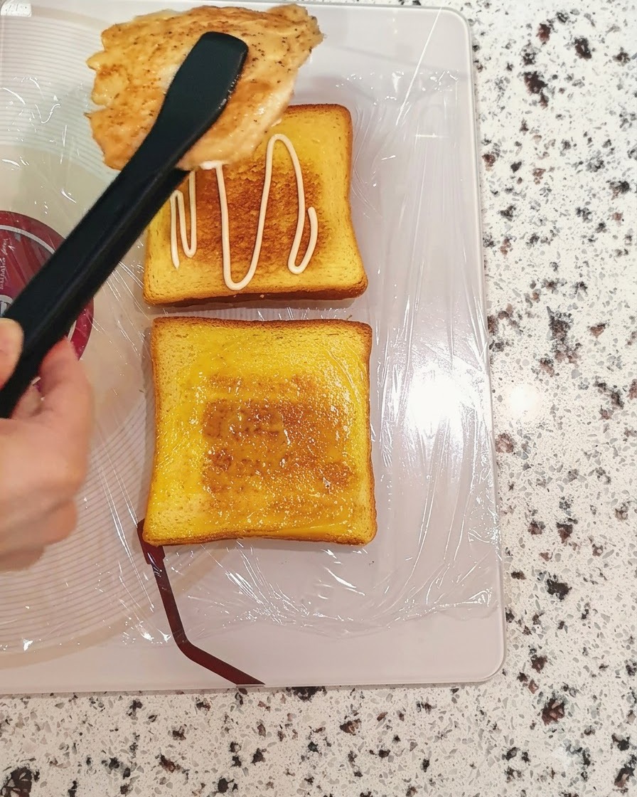 Gekochte Hähnchenbrust wird mit einer Zange auf eine mit Sauce bestrichene Toastscheibe gelegt.