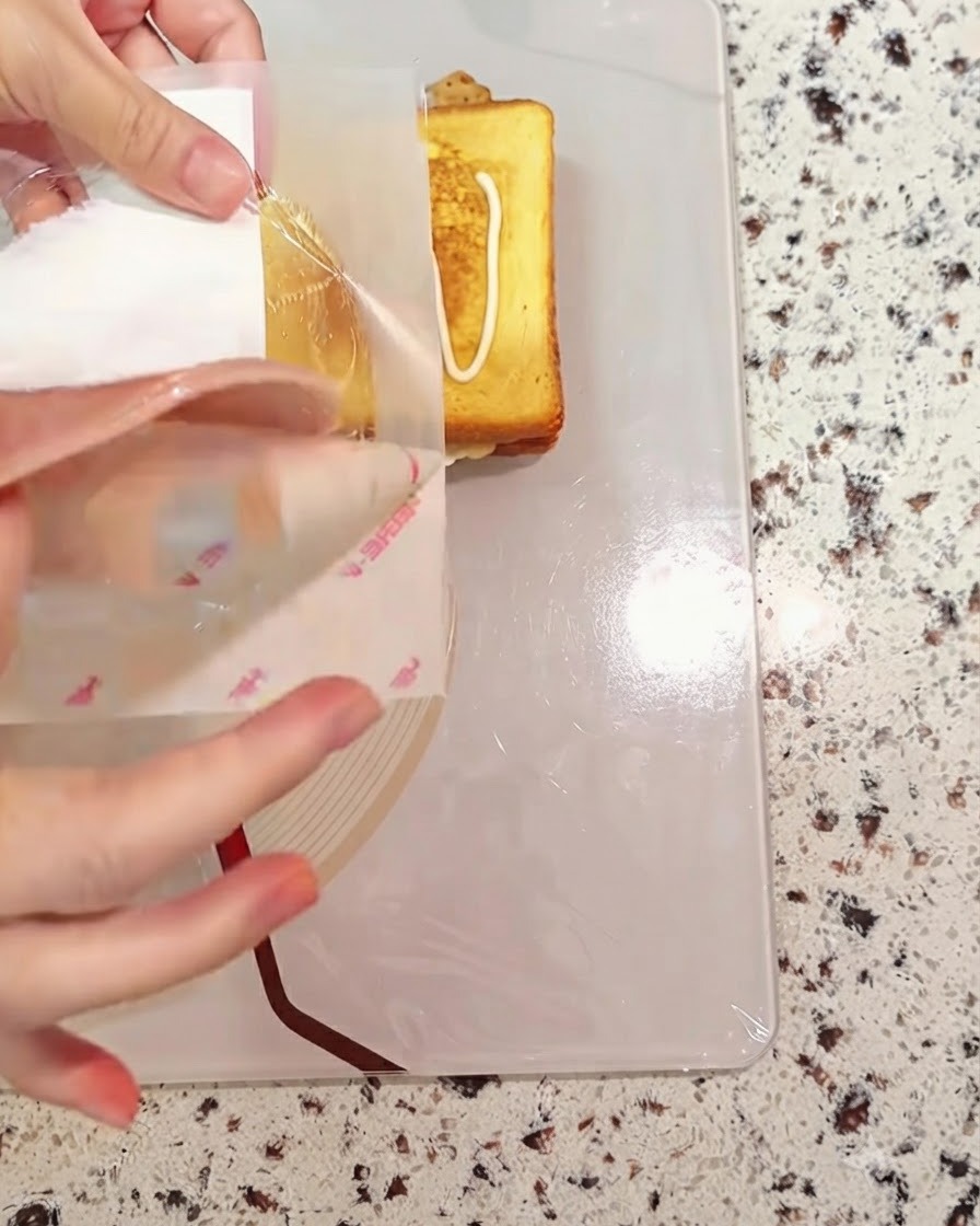 Eine Scheibe Schinken wird auf das sich im Aufbau befindliche Sandwichbrot gelegt.