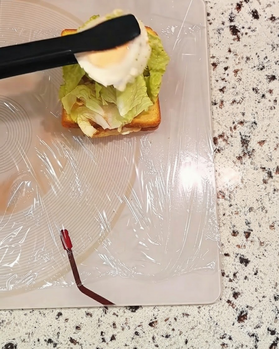 Eine schwarze Zange hält ein gekochtes Ei über eine Sandwichschicht mit Salat.