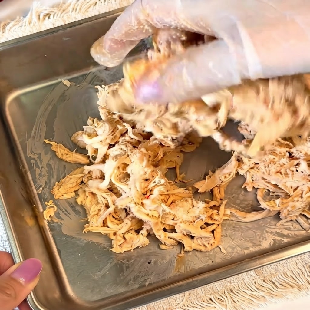 Hände mit Handschuhen mischen gewürztes, zerkleinertes Hähnchen in einem Metallbehälter.