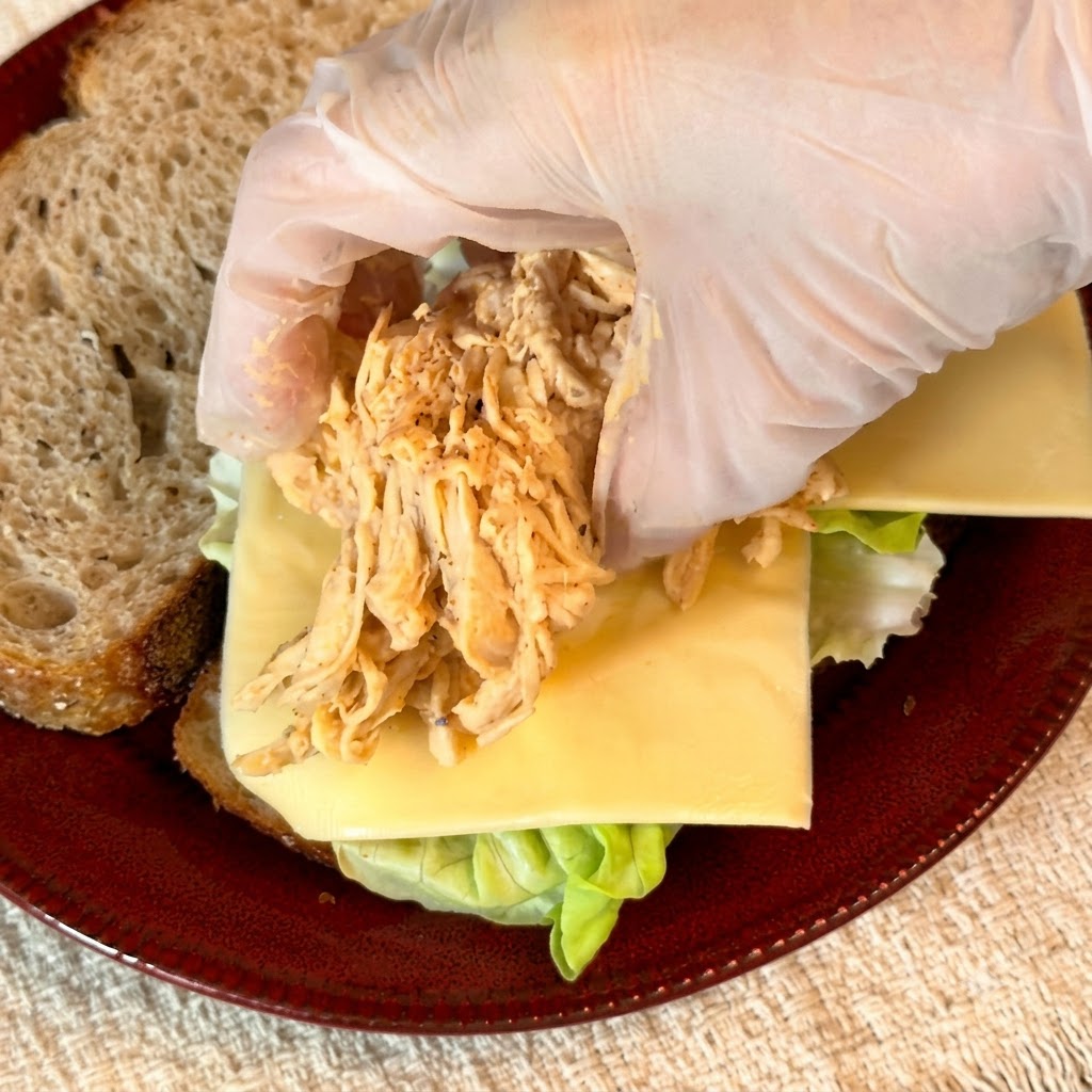 Handschuhe platzieren einen Berg gewürztes, zerkleinertes Hähnchen auf der Käseschicht des Sandwiches.
