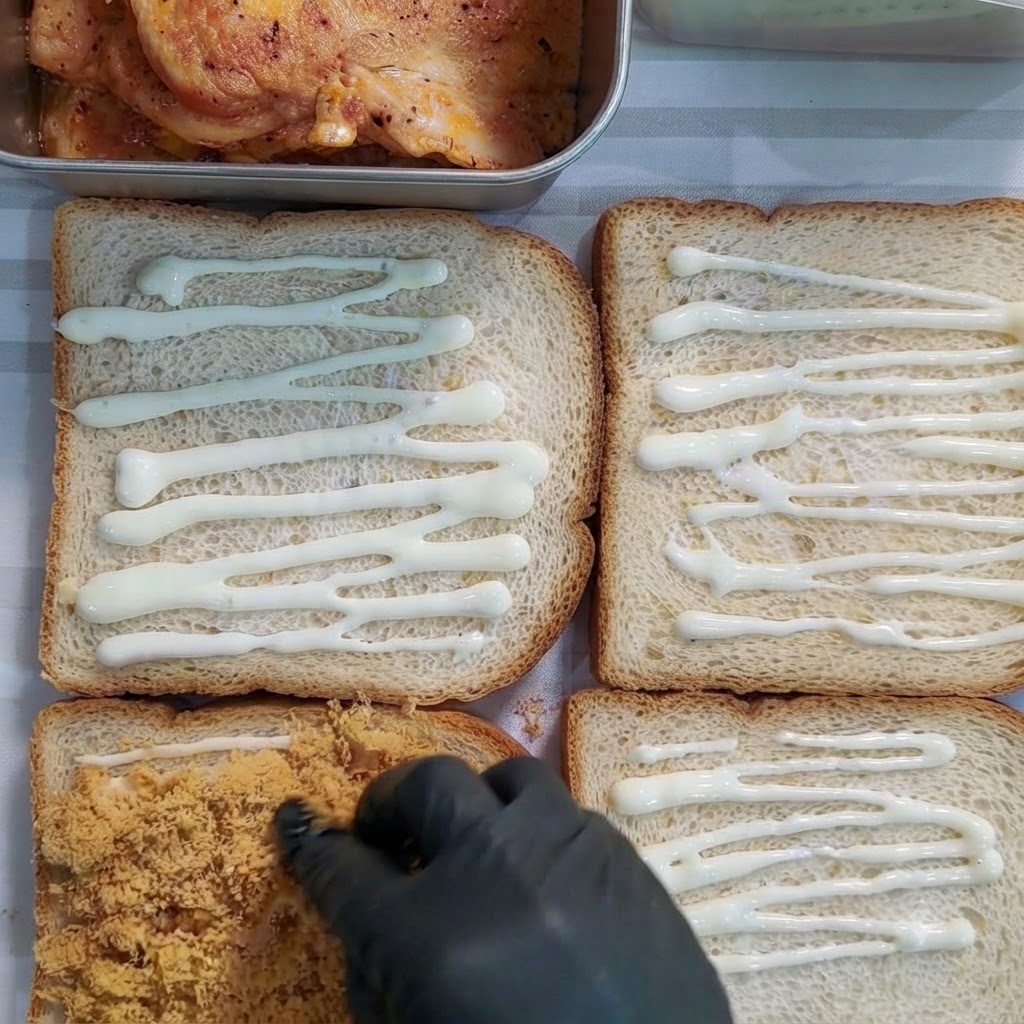 Eine behandschuhte Hand verteilt goldbraune Fleischflocken auf einer mit Dressing bedeckten Brotscheibe.