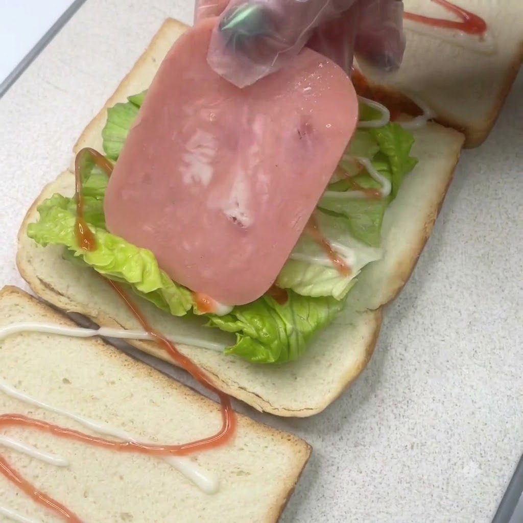 Eine Schinkenscheibe wird auf eine Salatschicht auf einem belegten Sandwichbrot mit Dressing gelegt.