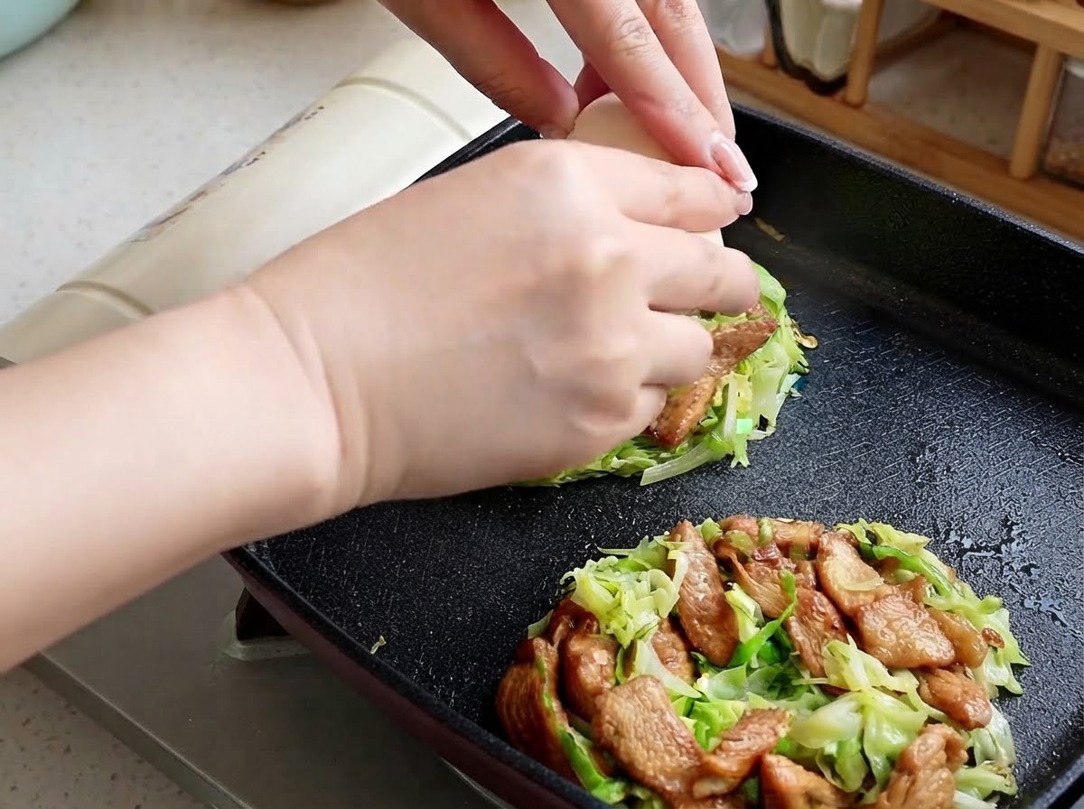 Hände schlagen ein rohes Ei auf die Mitte einer Kohl-Hähnchen-Portion in einer Pfanne.