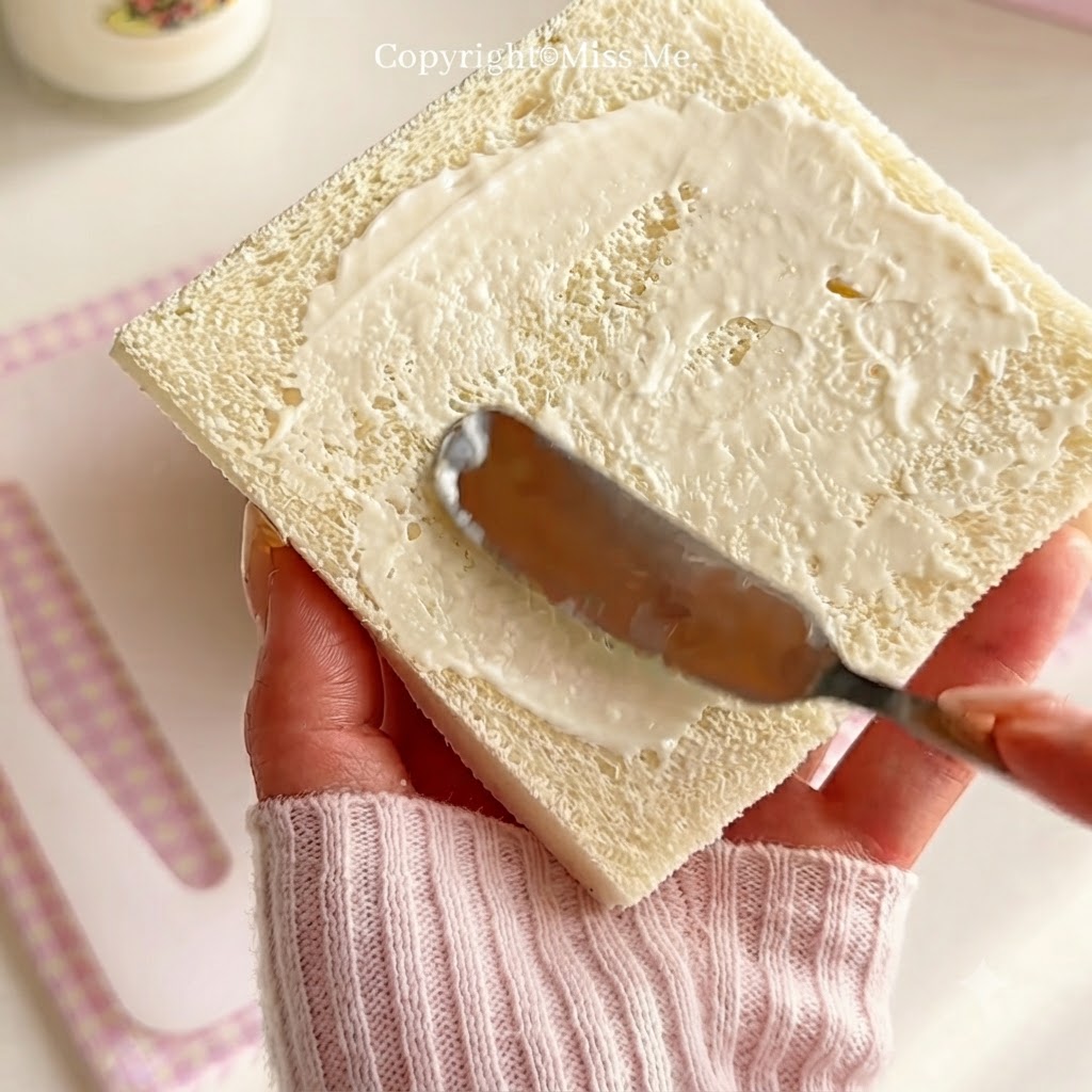 Eine Hand hält ein Messer, um weißes Salatdressing gleichmäßig über eine quadratische Weißbrotscheibe zu verteilen.
