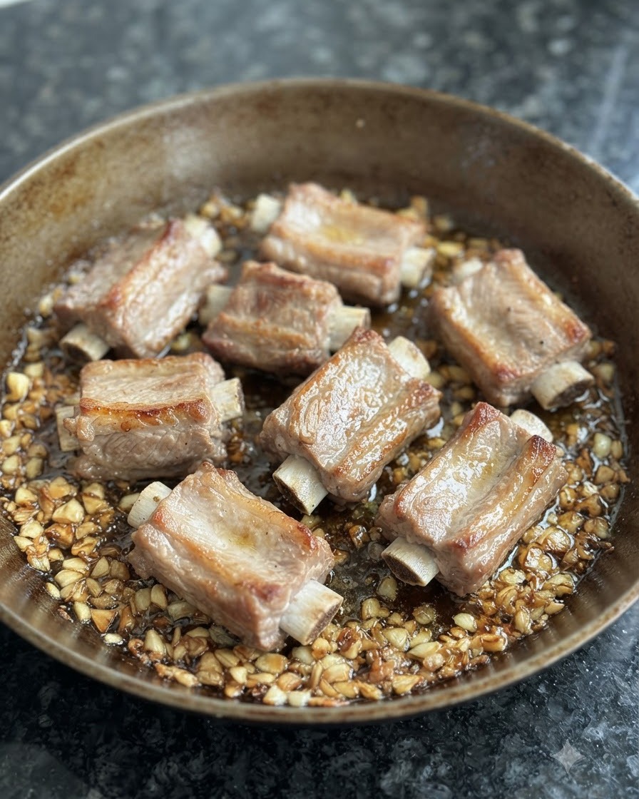 Blanchierte Schweinerippchen bräunen in einer Pfanne umgeben von gebratenem Knoblauch und Öl.
