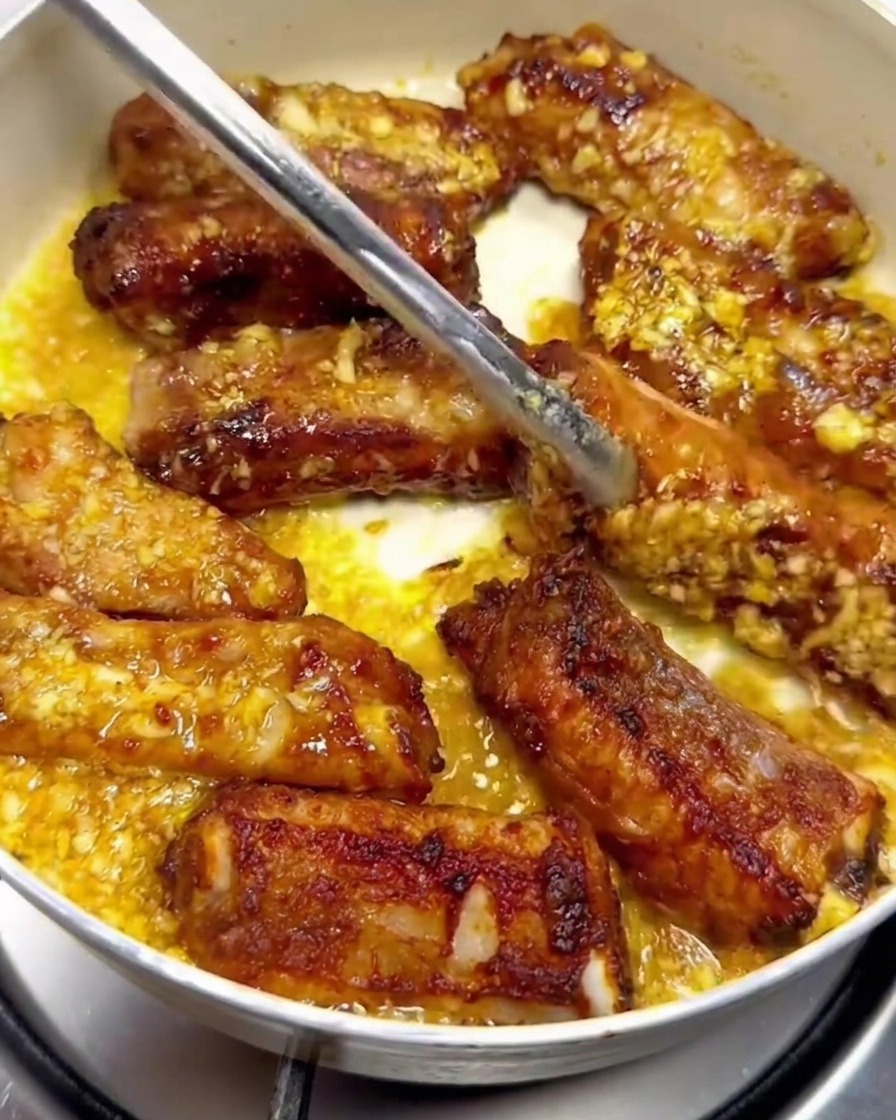 Schweinerippchen werden mit einer Metallzange in einer Pfanne gewendet und mit Knoblauch-Buttersauce überzogen.