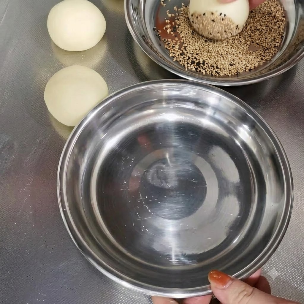 Ein Teigballen wird in einer Metallschüssel neben anderen vorbereiteten Teigballen in Sesam gewendet.