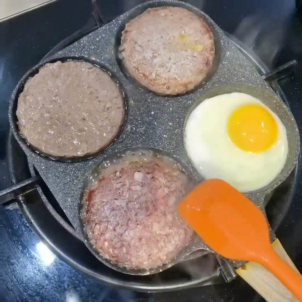 Burger-Patties werden mit einem orangefarbenen Spatel neben einem in einer Pfannensektion bratenem Ei gewendet.