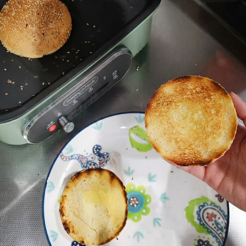 Getoastete Burgerbrötchenhälften auf einem Teller neben einer Pfanne mit einem ganzen Brötchen.