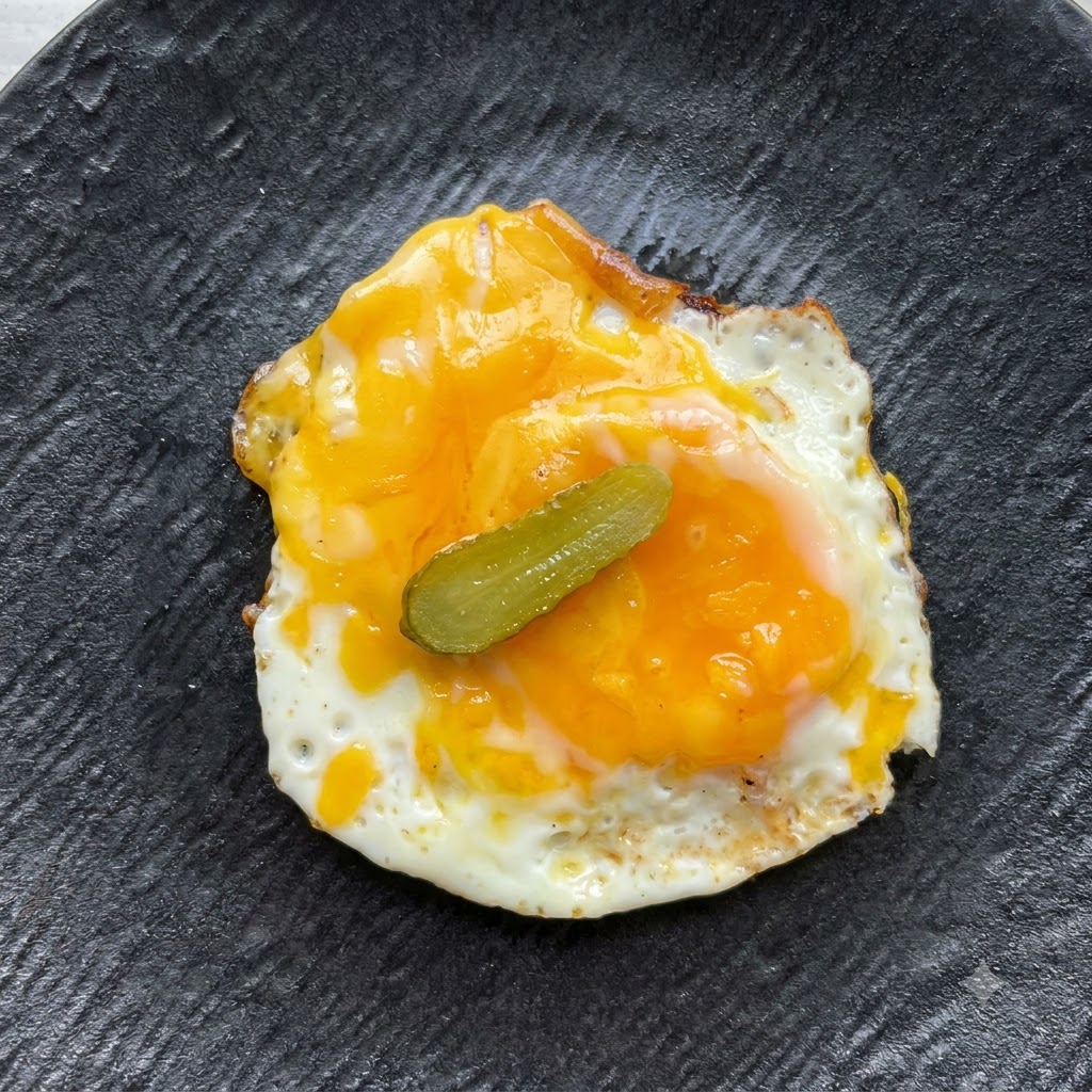 Eine einzelne kleine Gurkenscheibe liegt zentral auf dem Spiegelei und dem Käse.