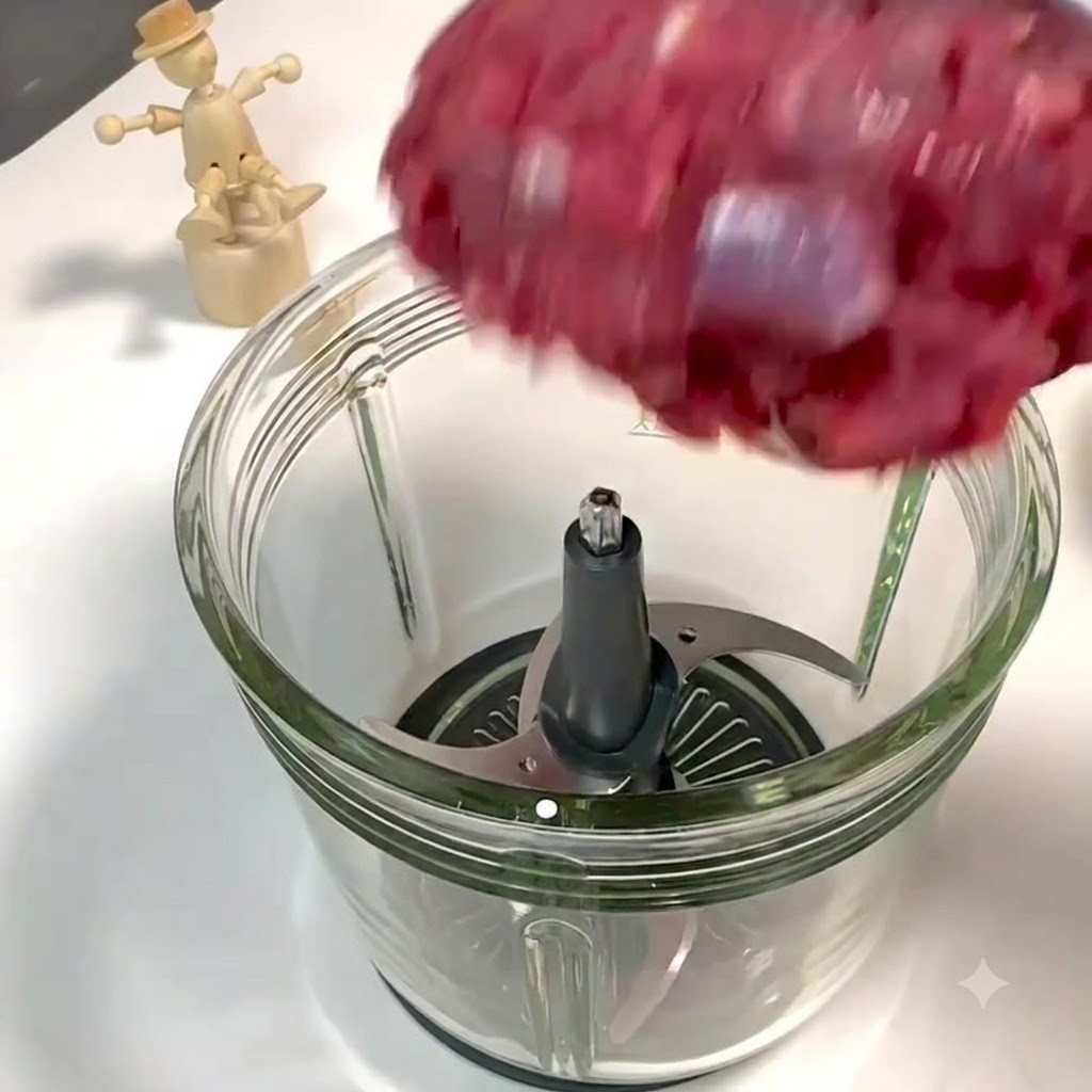Stücke von Rinderlende und Schweinebauch fallen in den offenen Glasbehälter einer Küchenmaschine über die Metallmesser.