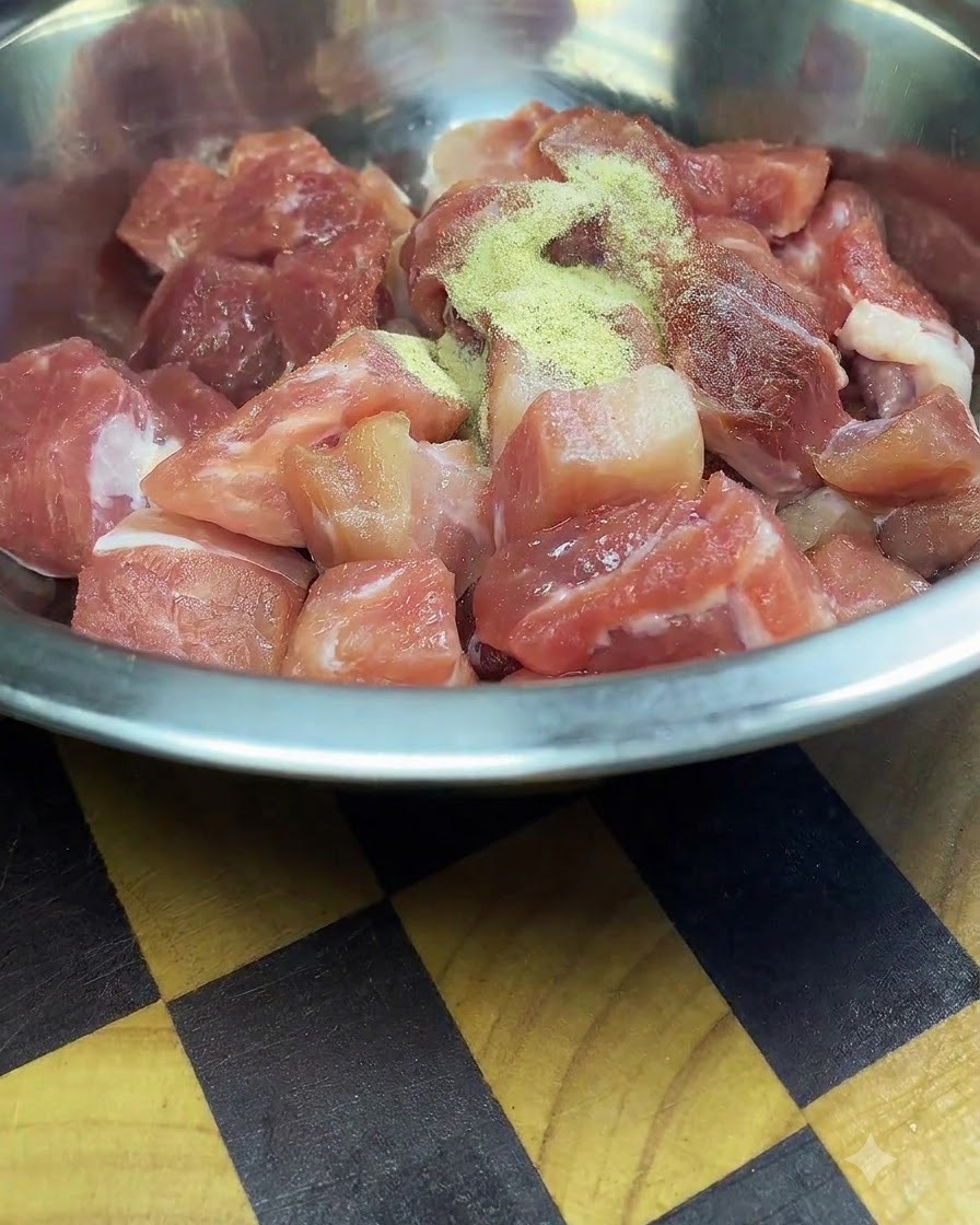 Frisch geschnittene rohe Schweinefleischwürfel in einer Edelstahlschüssel, bestreut mit Gewürzen und Maisstärke.