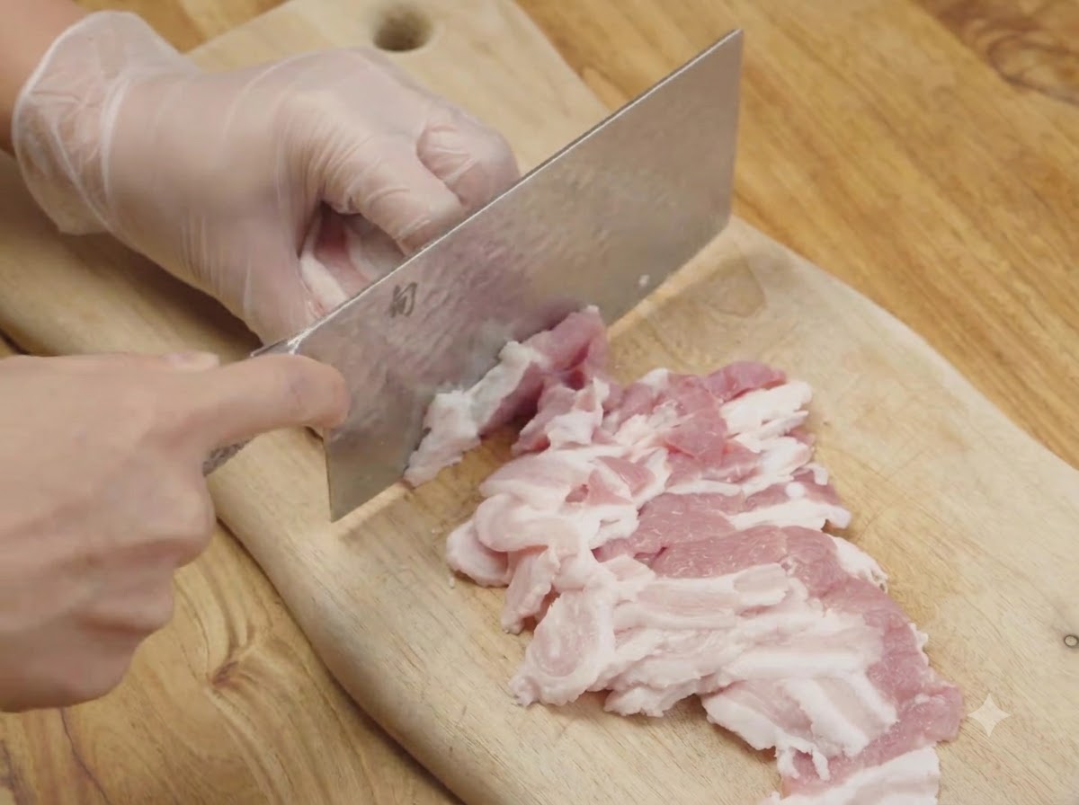 Roher Schweinebauch wird auf einem Holzschneidebrett mit einem Küchenbeil in dünne Streifen geschnitten.